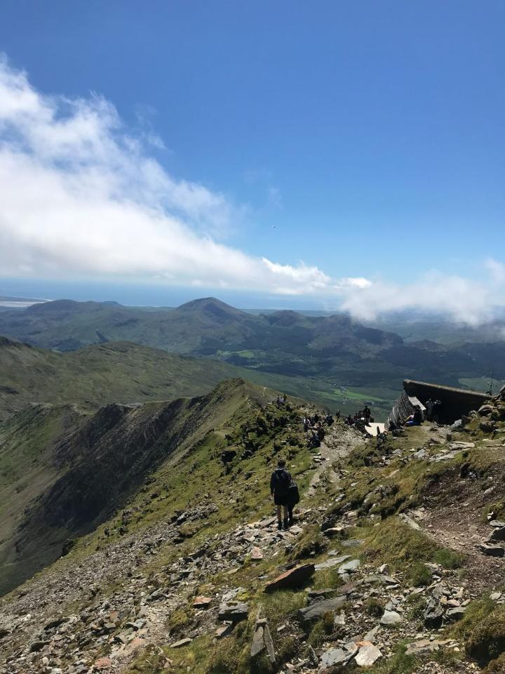 A walker on a rocky ridge near the top of Snowdon, with hills and the coast in the distance
